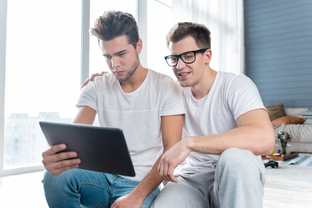 Uomini gay rilassati gioiosi che sorridono e utilizzano un tablet seduti vicini gli uni agli altri sul pavimento.