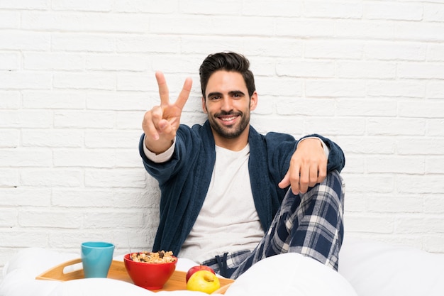 Uomo a letto con vestaglia e fare colazione sorridendo e mostrando il segno della vittoria