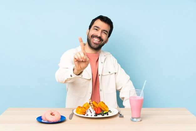 Uomo a un tavolo con cialde per la colazione e un frappè che mostra e alza un dito