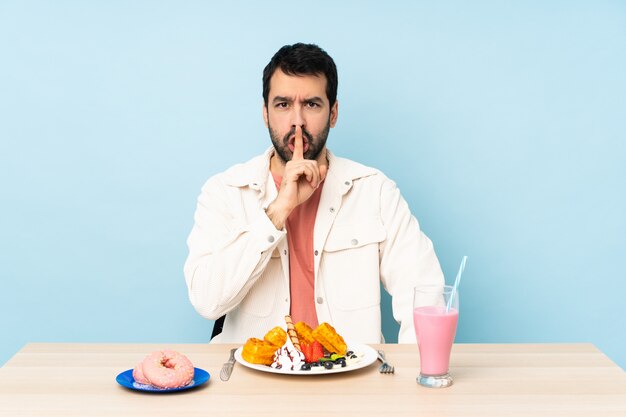 Uomo a un tavolo con cialde per la colazione e un frappè che mostra un segno di silenzio gesto mettendo il dito in bocca