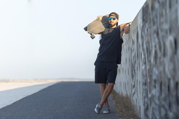 Uomo adulto attraente in piedi vicino al muro che tiene uno skateboard