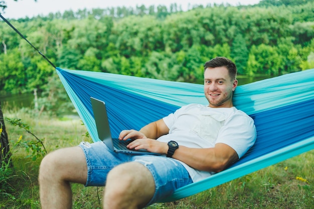 Uomo adulto bello che si rilassa in un'amaca mentre lavora al portatile durante l'escursione di giorno all'aperto Ricreazione in natura con lavoro a distanza