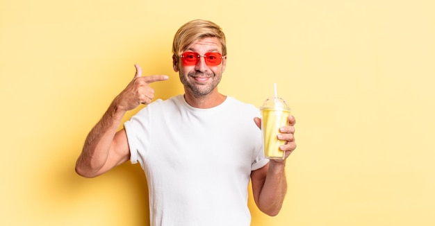 Uomo adulto biondo che sorride con sicurezza indicando il proprio ampio sorriso con un milkshake