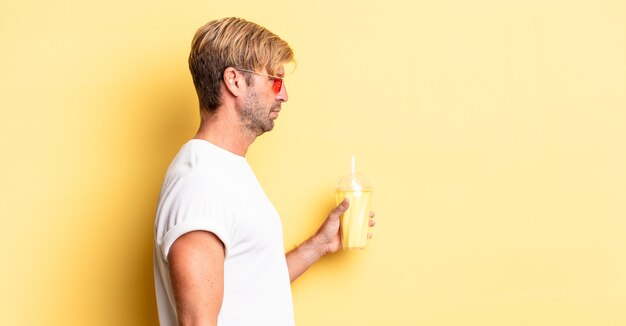 Uomo adulto biondo sulla vista di profilo pensando, immaginando o sognando ad occhi aperti con un milkshake