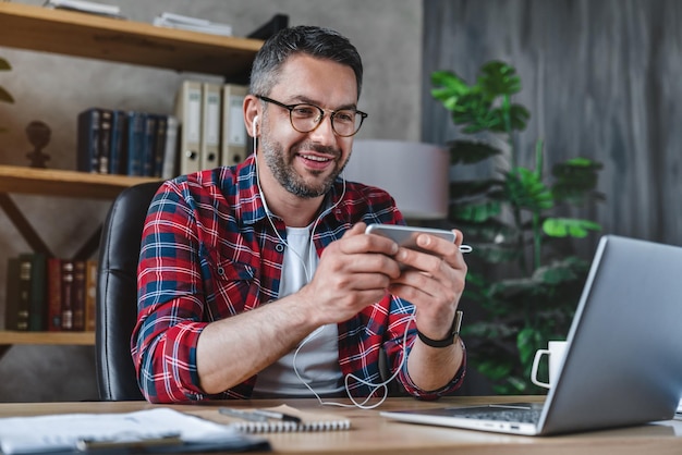 Uomo adulto che guarda video sul suo telefono con gli auricolari mentre usa il suo laptop nell'ufficio di casa