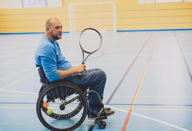Uomo adulto con disabilità fisica su sedia a rotelle che gioca a tennis su un campo da tennis al coperto