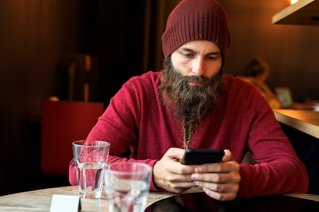 Uomo adulto con la barba intrecciata seduto al bar con il computer portatile che beve una tazza di caffè mentre digita sul telefono