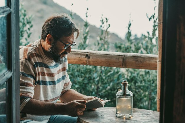 Uomo adulto maturo seduto a casa nella terrazza all'aperto a leggere un libro e rilassarsi godendosi la natura fuori sullo sfondo - i maschi rilassati amano leggere e studiare - ritratto barbuto dell'uomo
