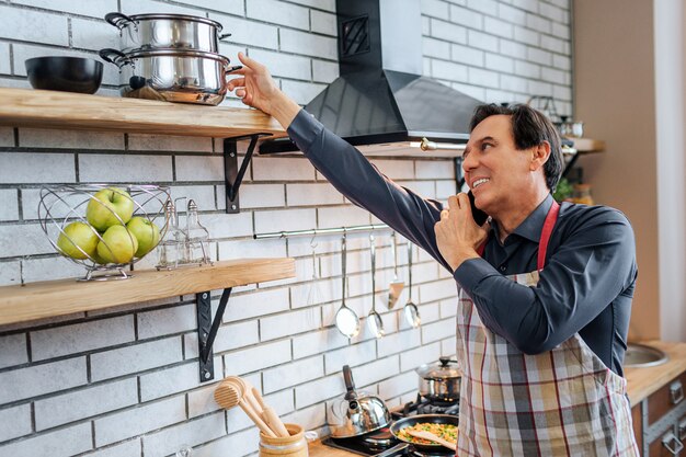 Uomo adulto stare in cucina e parlare al telefono. Allunga la mano per andare in padella. Guy indossare grembiule. È solo.