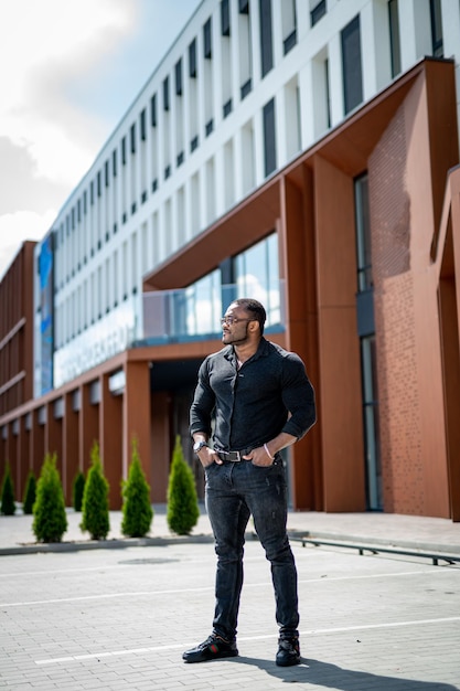 Uomo africano che indossa abiti scuri in piedi al business center Ragazzo sportivo in piedi con le mani nelle tasche dei jeans Foto a figura intera di un bell'uomo in strada