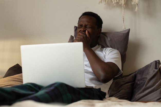 Uomo africano che lavora sdraiato sul letto a casa essendo stanco che sbadiglia