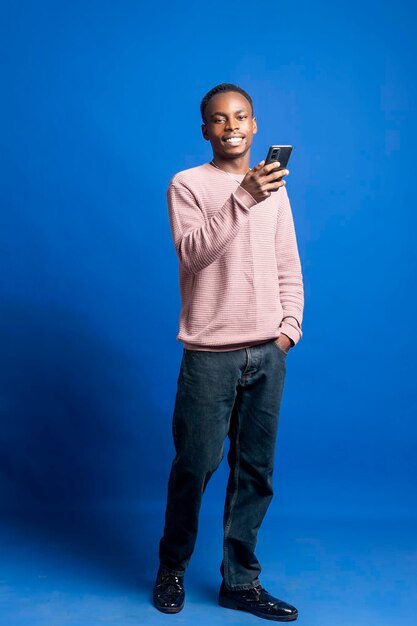 Uomo africano nero giovane e soddisfatto che usa uno smartphone su uno sfondo blu scuro pulito
