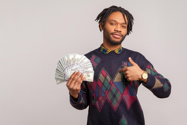 Uomo africano sicuro di sé soddisfatto con barba e dreadlocks che mostra i pollici in su gesto che tiene un fan di dollari accredito di cashback Indoor studio shot isolato su sfondo grigio