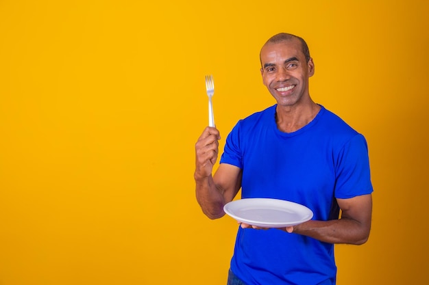 Uomo afro che tiene piatto vuoto e forchetta con isolato su sfondo giallo Copia spazio e mock up