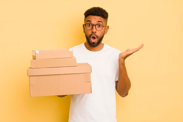 Uomo afro nero che sembra sorpreso e scioccato con la mascella caduta tenendo un concetto di scatole di spedizione di oggetti