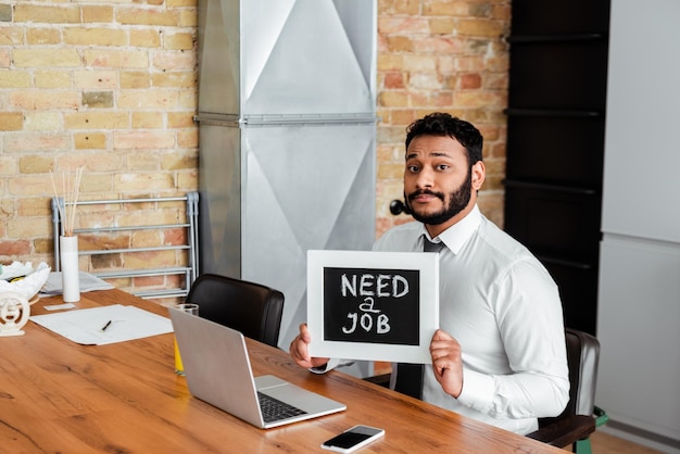 Uomo afroamericano barbuto che tiene la lavagna con bisogno di un lavoro scritto vicino ai gadget