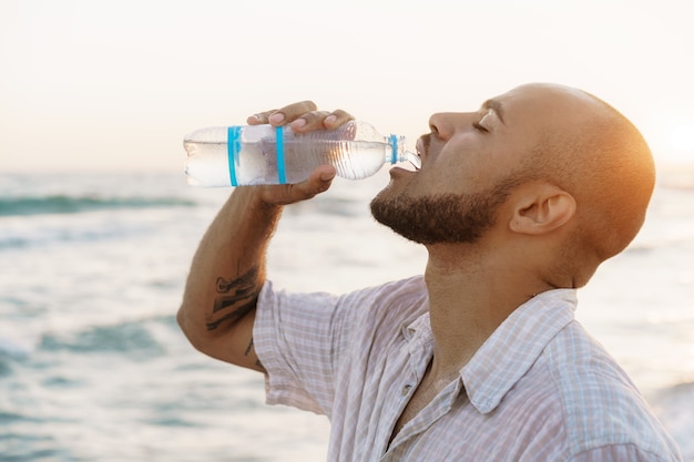 Uomo afroamericano che beve acqua dalla bottiglia sulla spiaggia