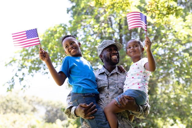 Uomo afroamericano che indossa un'uniforme militare, torna a casa, in piedi in un giardino, tiene e abbraccia i suoi figli, i bambini tengono e sventolano mini bandiere