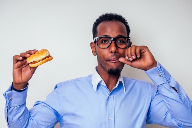 Uomo afroamericano che si gode il gusto dell'hamburger. bello e giovane uomo afro in una camicia elegante e occhiali che tengono un hamburger su uno sfondo bianco. amore per la dieta del cibo spazzatura