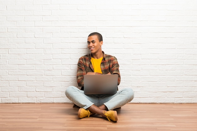 Uomo afroamericano che si siede sul pavimento con il suo computer portatile che tiene le armi attraversate nella posizione laterale mentre sorridendo