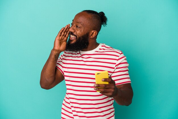 Uomo afroamericano che tiene un telefono cellulare isolato sulla parete blu che grida e che tiene il palmo vicino alla bocca aperta.