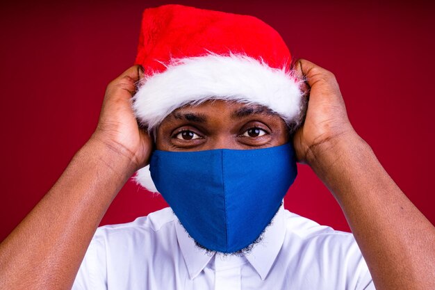 Uomo afroamericano con cappello da Babbo Natale che indossa una maschera sullo sfondo rosso dello studio