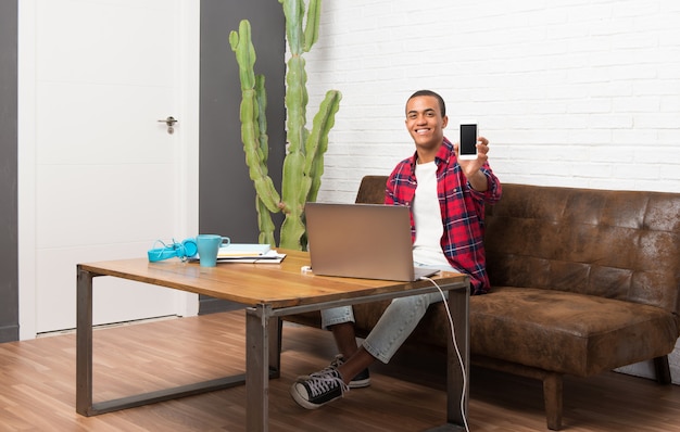 Uomo afroamericano con il computer portatile nel salone che esamina la macchina fotografica e che sorride mentre usando il cellulare