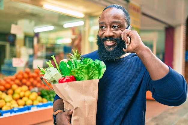 Uomo afroamericano con la barba che tiene un sacchetto di carta di generi alimentari dal supermercato che parla al telefono