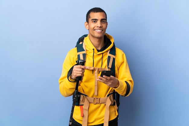 Uomo afroamericano con zaino e bastoncini da trekking su sfondo isolato inviando un messaggio con il cellulare