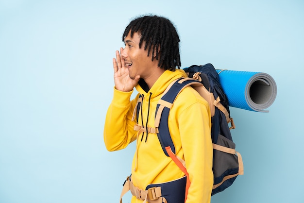 Uomo afroamericano del giovane alpinista con un grande zaino isolato su una parete blu che grida con la bocca spalancata