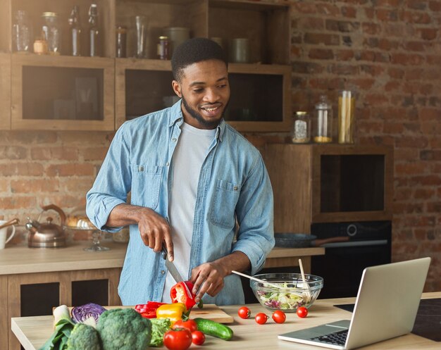 Uomo afroamericano felice che prepara insalata di verdure mentre guarda sul laptop