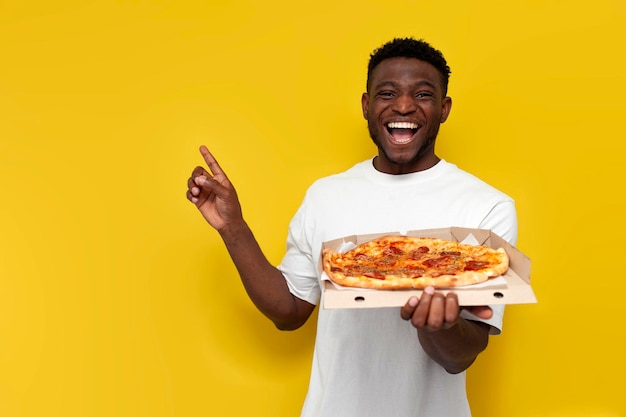 uomo afroamericano gioioso in maglietta bianca tiene in mano una scatola di pizza e indica con la mano sul lato