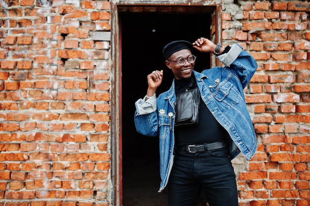 Uomo afroamericano in giacca di jeans berretto e occhiali contro il muro di mattoni al tetto abbandonato