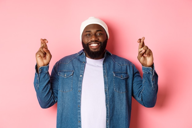 Uomo afroamericano ottimista che esprime desiderio, tenendo le dita incrociate e sorridendo, in piedi su sfondo rosa