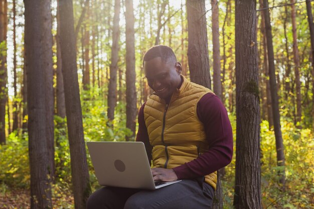 Uomo afroamericano seduto nel parco cittadino su una panchina con il computer portatile che studia online all'aperto uomo freela