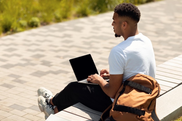 Uomo afroamericano seduto su una panchina e lavorando online su un computer portatile con schermo vuoto dentro