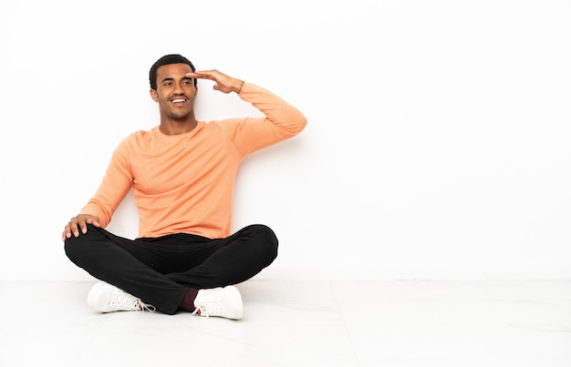 Uomo afroamericano seduto sul pavimento su sfondo copyspace isolato guardando lontano con la mano per guardare qualcosa