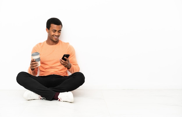 Uomo afroamericano seduto sul pavimento su sfondo copyspace isolato tenendo il caffè da portare via e un cellulare