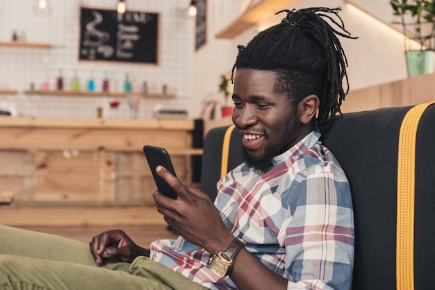 uomo afroamericano sorridente che usa uno smartphone in un caffè