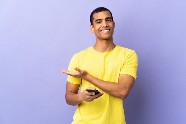 Uomo afroamericano su sfondo viola isolato utilizzando il telefono cellulare che estende le mani a lato per invitare a venire