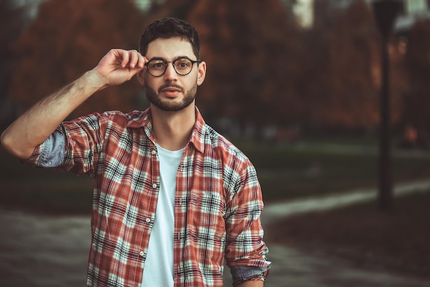 Uomo alla moda con la barba e occhiali da sole in piedi nel parco.