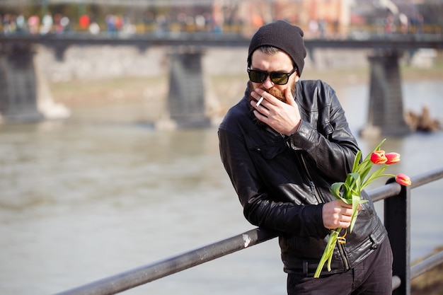 Uomo alla moda con la barba in occhiali da sole con i baffi in un caldo cappello e giacca di pelle tiene un mazzo di tulipani rossi e fuma una sigaretta