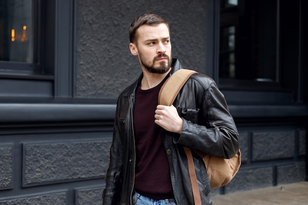 Uomo alla moda con la barba in piedi per strada con un cappotto di pelle. Servizio fotografico uomini per strada. Un uomo in uno stile alla moda. Acconciatura da uomo elegante