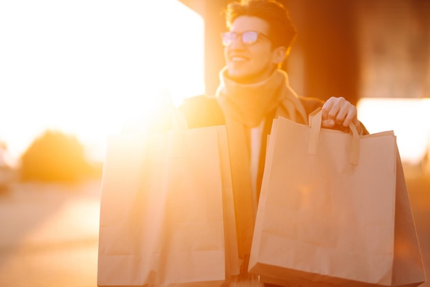 Uomo alla moda con pacchetti di carta dopo lo shopping al tramonto Uomo bello che fa shopping