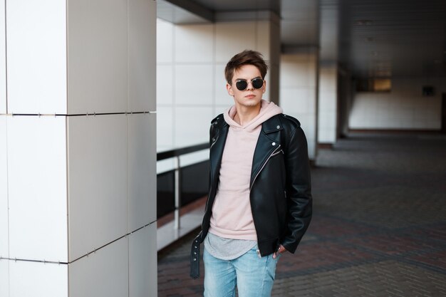 Uomo alla moda giovane hipster in occhiali da sole neri in elegante giacca nera in felpa rosa in jeans alla moda in una t-shirt grigia è in piedi al chiuso. Ragazzo moderno con un'acconciatura alla moda.