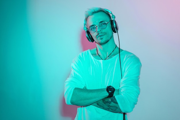 Uomo alla moda hipster in eleganti occhiali rotondi che ascolta musica in studio con luce blu e rosa creativa