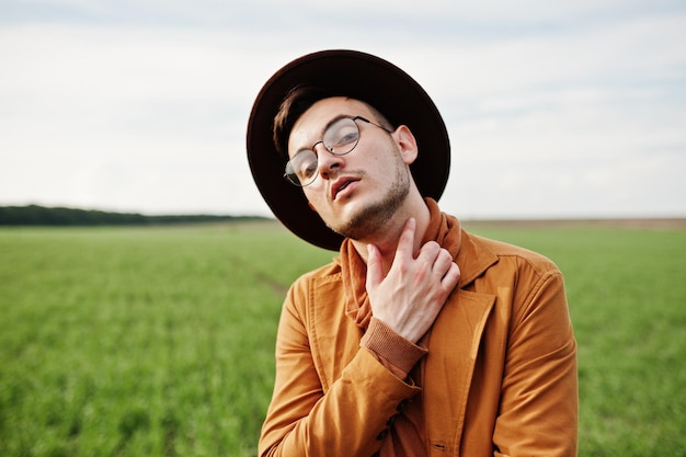 Uomo alla moda in giacca marrone occhiali e cappello posato su campo verde