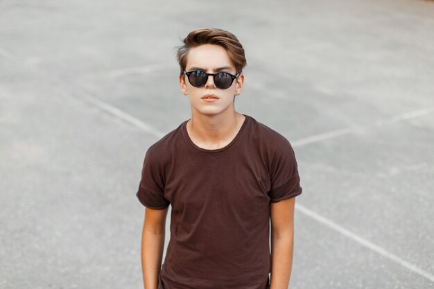 Uomo alla moda urbano giovane hipster in elegante maglietta marrone con acconciatura in occhiali da sole neri alla moda si trova allo stadio