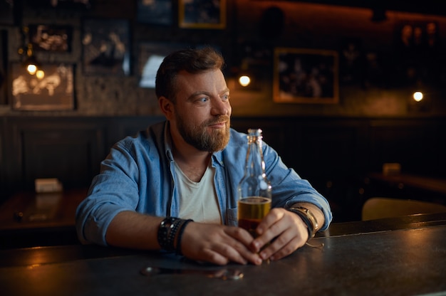 Uomo allegro con una bottiglia di birra seduto al bancone del bar