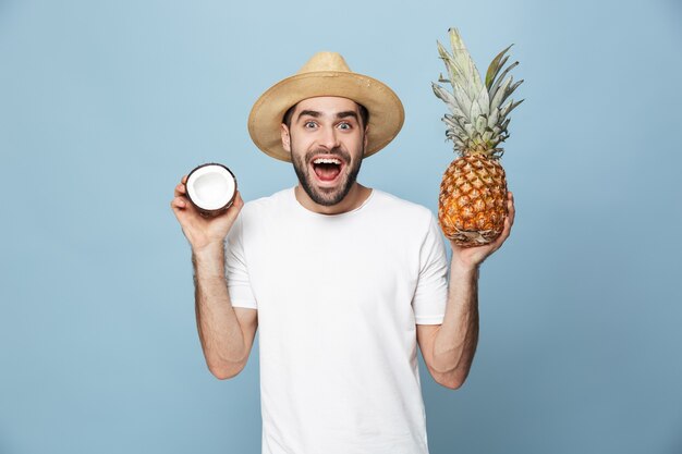 Uomo allegro eccitato che indossa una maglietta bianca in piedi isolato su un muro blu, con in mano ananas e cocco